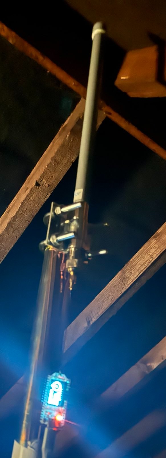 An antenna mounted on a metal pole attached to a wooden beam with a visible electronics module displaying blue and red illuminated indicators.